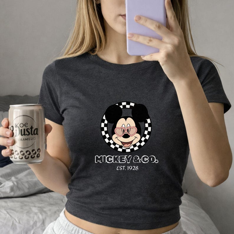 Puede incluir: Camiseta gris oscuro con un dise&ntilde;o de Mickey Mouse con gafas de sol en forma de coraz&oacute;n, enmarcado por un c&iacute;rculo a cuadros. El texto "MICKEY & CO. EST. 1928" est&aacute; impreso debajo. Una persona sostiene una lata de refresco.