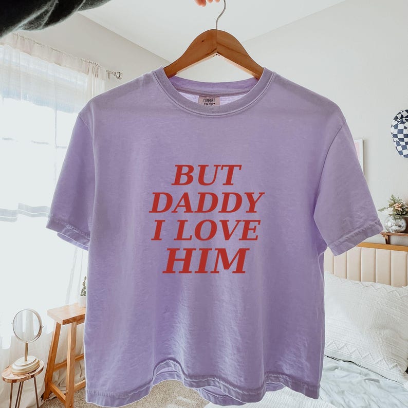 May include: A light purple short-sleeved t-shirt featuring the red text "BUT DADDY I LOVE HIM" printed in a bold, serif font across the center of the chest. The garment has a crew neckline and is displayed on a wooden hanger against a bright room.