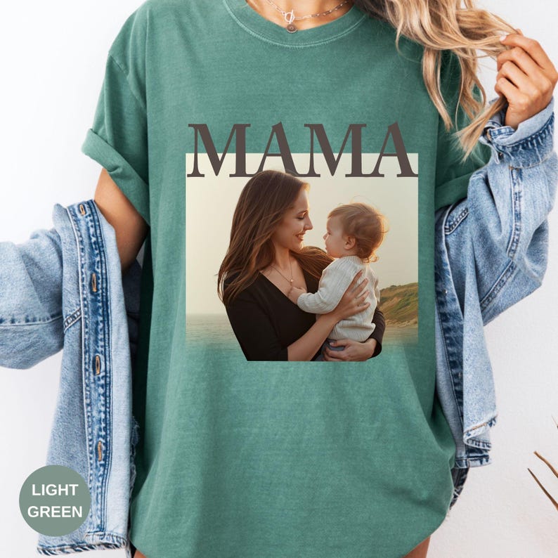 May include: Light green t-shirt with the word "MAMA" in dark gray block letters above a framed photograph of a mother and child. The photograph shows a woman holding a baby, with a scenic background.