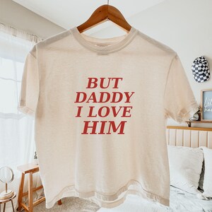 May include: A cream-colored short-sleeve t-shirt featuring the red text "BUT DADDY I LOVE HIM" printed in a bold, serif font across the center of the chest. The garment is displayed on a wooden hanger against a neutral indoor setting.