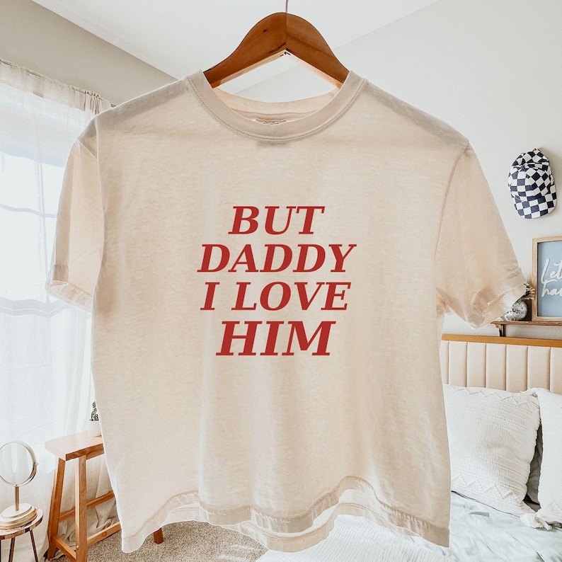 May include: A cream-colored short-sleeve t-shirt featuring the red text "BUT DADDY I LOVE HIM" printed in a bold, serif font across the center of the chest. The garment is displayed on a wooden hanger against a neutral indoor setting.