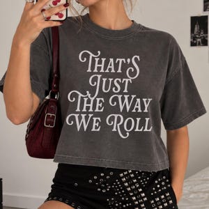 May include: A charcoal grey short-sleeve crop top featuring the white text That's Just The Way We Roll in a decorative serif font. The garment has a crew neckline and a boxy fit, paired with a dark red shoulder bag and black shorts.