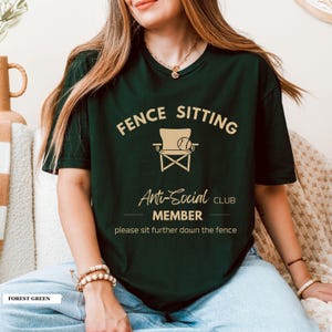 May include: Dark green t-shirt featuring the text "FENCE SITTING Anti-Social CLUB MEMBER" and a chair and tennis ball graphic. The shirt also includes the phrase "please sit further down the fence".
