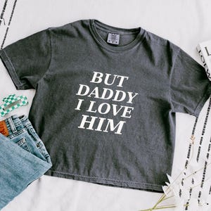 May include: A charcoal gray short-sleeve t-shirt with the white text "BUT DADDY I LOVE HIM" printed on the front. The shirt features a crew neckline and a relaxed fit, displayed alongside a pair of blue denim jeans and a small green checkered accessory.