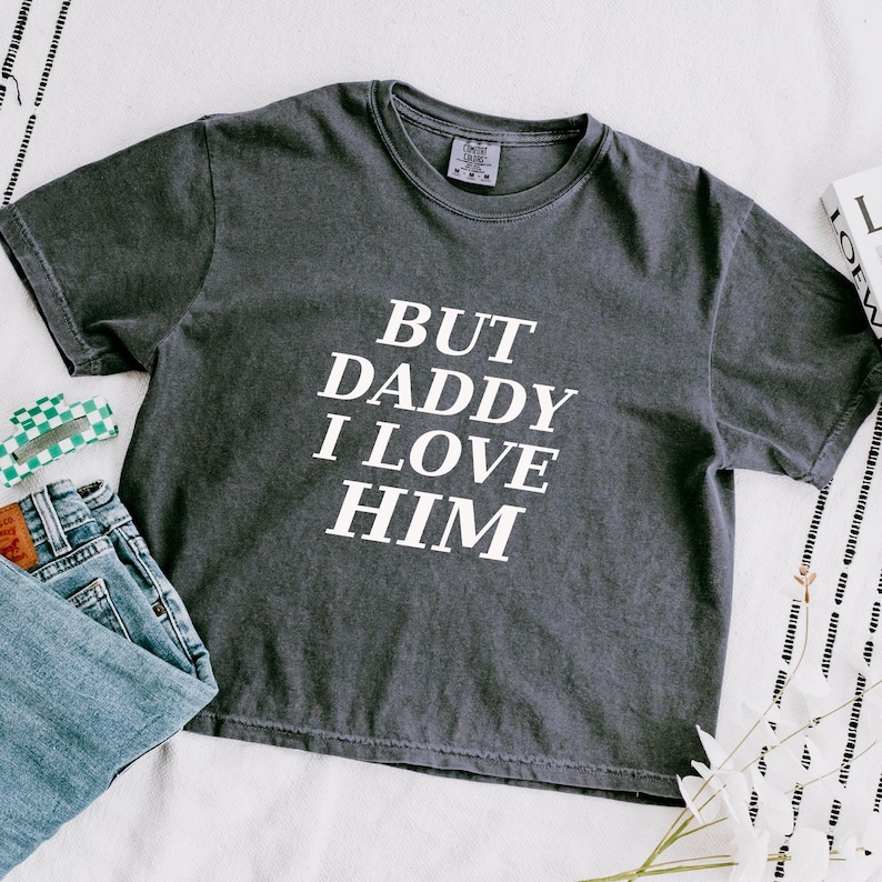 May include: A charcoal gray short-sleeve t-shirt with the white text "BUT DADDY I LOVE HIM" printed on the front. The shirt features a crew neckline and a relaxed fit, displayed alongside a pair of blue denim jeans and a small green checkered accessory.