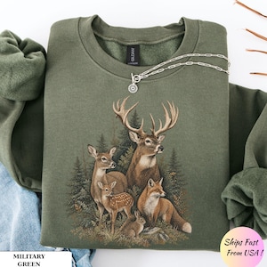 May include: A military green crewneck sweatshirt featuring a detailed graphic print of a buck, a doe, a fawn, a fox, and a rabbit in a forest setting. The garment has a Gildan brand label at the inner neckline and a silver-toned chain necklace.
