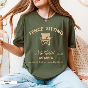 May include: A military green t-shirt featuring the words "FENCE SITTING Anti-Social CLUB MEMBER" and a chair and ball graphic. The text "please sit further down the fence" is printed below. The shirt is a solid colour.