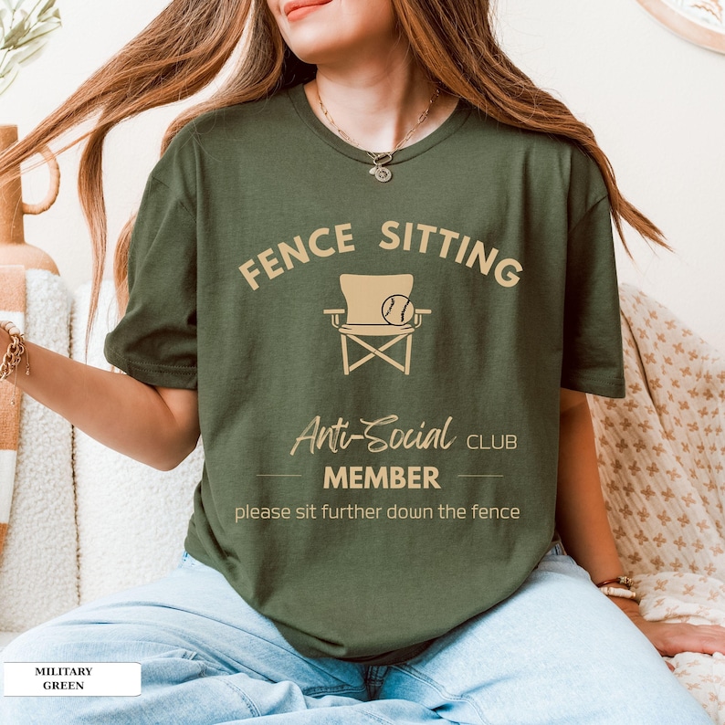May include: A military green t-shirt featuring the words "FENCE SITTING Anti-Social CLUB MEMBER" and a chair and ball graphic. The text "please sit further down the fence" is printed below. The shirt is a solid colour.