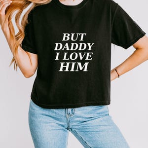 May include: A black short-sleeved t-shirt featuring the white text BUT DADDY I LOVE HIM printed in a bold, serif font across the chest. The garment has a crew neckline and a relaxed fit, paired with light blue denim jeans.