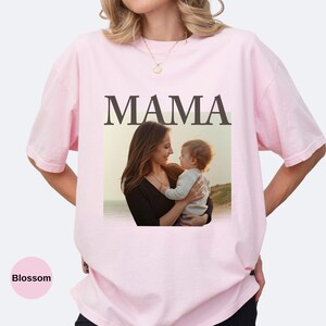 May include: A light pink t-shirt with the word "MAMA" in large, dark brown letters. Below the word is a photo of a mother holding a baby. The shirt is a soft, comfortable material.