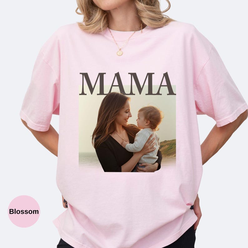 May include: A light pink t-shirt with the word "MAMA" in large, dark brown letters. Below the word is a photo of a mother holding a baby. The shirt is a soft, comfortable material.