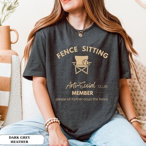May include: Dark grey heather t-shirt with the text "FENCE SITTING Anti-Social CLUB MEMBER" and a graphic of a chair and a ball. The shirt also includes the phrase "please sit further down the fence".
