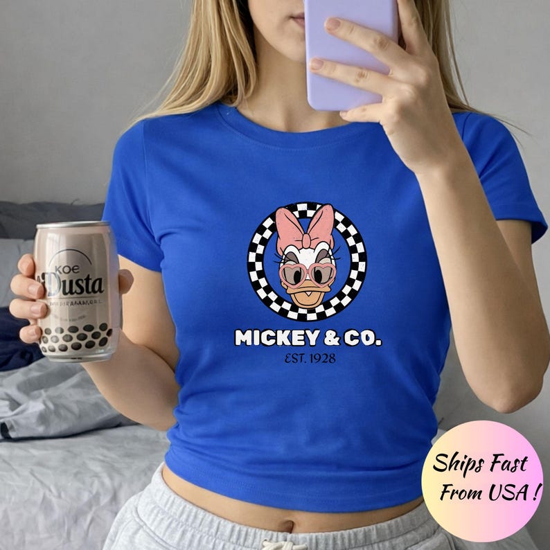 Puede incluir: Camiseta azul real con un gr&aacute;fico de Daisy Duck, lazo rosa, gafas y un dise&ntilde;o de c&iacute;rculo a cuadros. El texto "MICKEY & CO. EST. 1928" est&aacute; debajo. En la imagen se sostiene una lata de Koe Dusta.