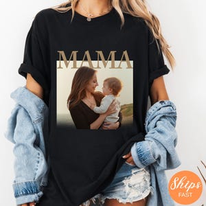 May include: Black t-shirt with the word "MAMA" in a gold-colored font. The shirt features a photo of a mother and child. The mother is looking at the child, who is being held in her arms. The background of the photo is a beach.