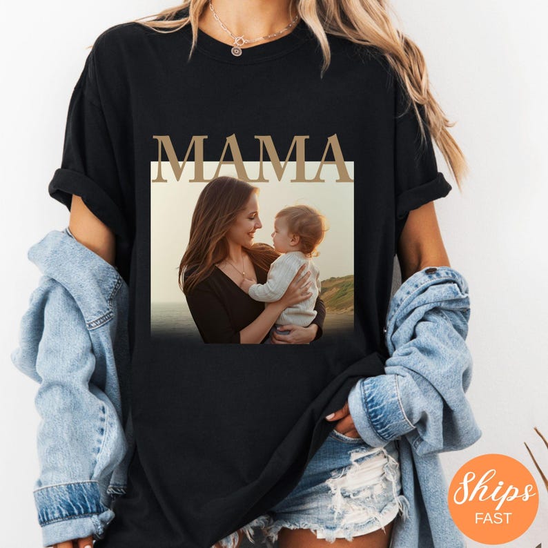 May include: Black t-shirt with the word "MAMA" in a gold-colored font. The shirt features a photo of a mother and child. The mother is looking at the child, who is being held in her arms. The background of the photo is a beach.