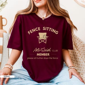 May include: A maroon t-shirt featuring the text "FENCE SITTING Anti-Social CLUB MEMBER" and a chair and baseball graphic. The shirt also includes the phrase "please sit further down the fence".