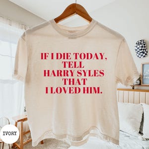 Puede incluir: Una camiseta de manga corta color marfil con texto rojo impreso en la parte delantera que dice: IF I DIE TODAY, TELL HARRY SYLES THAT I LOVED HIM. La camiseta se muestra en una percha de madera sobre un fondo claro.