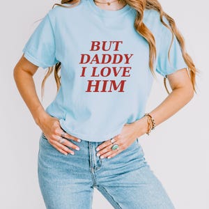 May include: A light blue short-sleeved t-shirt featuring the text BUT DADDY I LOVE HIM printed in a bold, dark red serif font across the chest. The garment has a classic crew neckline and a standard fit, paired with blue denim jeans.