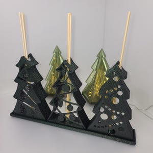 Può includere: Un'esposizione decorativa di alberi di Natale con tre ritagli a forma di albero verde scuro e glitterato con tasselli di legno. Dietro ci sono due luci traslucide a forma di albero verde. L'esposizione è progettata per contenere i tasselli.