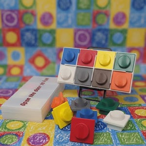 May include: A rectangular white plastic case with the text Open the door to Fun printed on the lid. Next to it is a small black metal stand holding a grid of eight colorful square plastic tiles, each with a central circular knob. Additional loose tiles in red, yellow, blue, gray, green, and white are scattered in front.