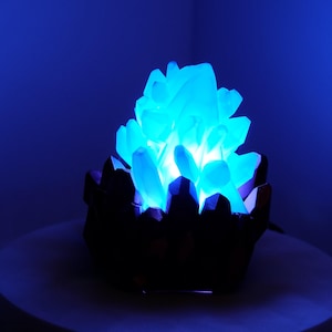 May include: A glowing blue crystal sculpture. The crystal structure is illuminated from within, creating a bright, ethereal glow. The base is dark, contrasting with the bright blue light. The overall effect is a striking, decorative piece.