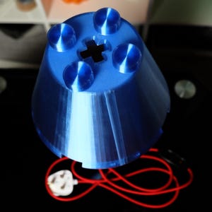 Peut inclure: Un abat-jour bleu de forme conique imprim&eacute; en 3D, dot&eacute; d'une surface sup&eacute;rieure avec quatre protub&eacute;rances circulaires et une d&eacute;coupe centrale en forme de croix. L'objet est reli&eacute; &agrave; un fin cordon &eacute;lectrique rouge avec une prise blanche.