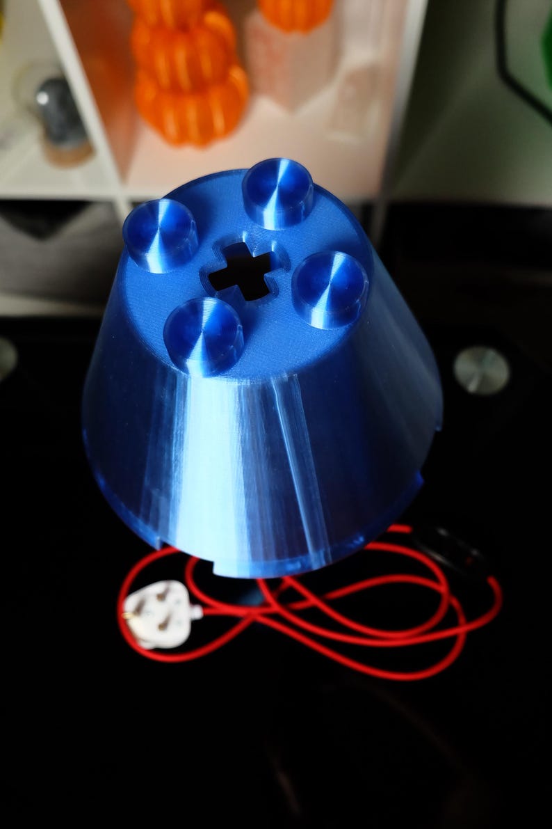 Peut inclure: Un abat-jour bleu de forme conique imprim&eacute; en 3D, dot&eacute; d'une surface sup&eacute;rieure avec quatre protub&eacute;rances circulaires et une d&eacute;coupe centrale en forme de croix. L'objet est reli&eacute; &agrave; un fin cordon &eacute;lectrique rouge avec une prise blanche.