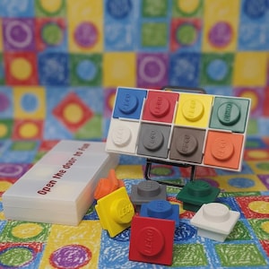 May include: A collection of colorful LEGO bricks, including red, yellow, blue, and green, are displayed. A white container with the text "Open the door to Fun" is also present. The background features a vibrant, patterned fabric.