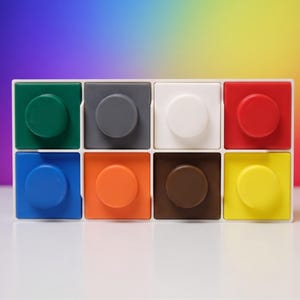 Pode incluir: Um organizador de armazenamento retangular branco com oito compartimentos quadrados dispostos em duas filas de quatro. Cada compartimento contém um bloco quadrado com um pino circular elevado no topo, nas cores verde, cinza, branco, vermelho, azul, laranja, marrom e amarelo.