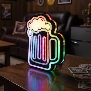 Peut inclure: Lampe LED en forme de chope de bière, avec un contour noir et un arc-en-ciel de couleurs à l'intérieur. Le haut de la chope est blanc, ressemblant à de la mousse. La lampe est sur une table en bois, avec des dessous de verre à proximité. C'est un objet décoratif.
