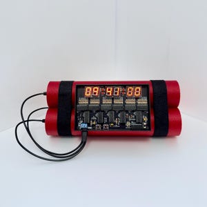 Puede incluir: Un dispositivo electrónico rojo que se asemeja a un cartucho de dinamita. Cuenta con pantallas digitales rojas, una placa de circuito negro y correas negras. Cables negros se extienden desde el dispositivo.