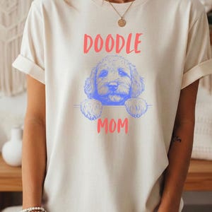 Op de afbeelding: Een crèmekleurig t-shirt met korte mouwen en een blauwe lijntekening van een hondengezicht en pootjes. De tekst "DOODLE" staat in het rood boven de hond en het woord "MOM" is in het rood onder de hondenillustratie op de borst gedrukt.