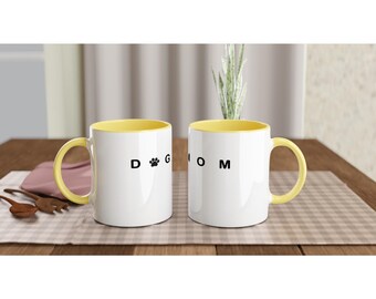 Puede incluir: Dos tazas de cerámica blancas con interior y asas amarillas. Una taza tiene el texto "DOG" con una huella de pata, y la otra dice "MOM" en letras negras. Las tazas están sobre una superficie a cuadros.