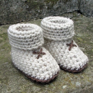 May include: A pair of beige crocheted baby booties with brown accents. The booties have a fold-over cuff and a simple cross design.