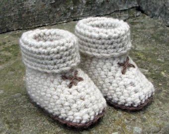 Pode incluir: Um par de sapatinhos de bebé em crochet bege com detalhes castanhos. Os sapatinhos têm um punho dobrado e um design simples de cruz.