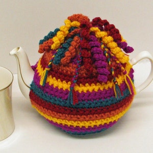 May include: A colorful crocheted teapot cozy with a striped pattern and tassels. The cozy is made in shades of red, orange, yellow, green, and purple.