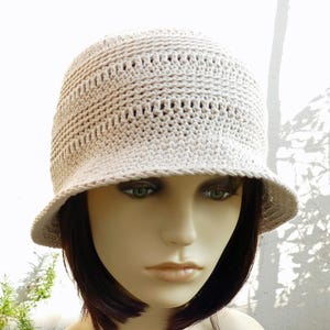 May include: A beige crochet bucket hat with a small brim. The hat has a textured appearance with small holes throughout the crown. The hat is displayed on a mannequin head with dark hair and green eyes. The hat is a neutral color.