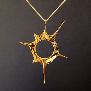 Op de afbeelding: Een gouden ketting met een hanger in de vorm van een stervormig ontwerp. De hanger heeft een ronde opening in het midden en is bevestigd aan een fijne gouden ketting.