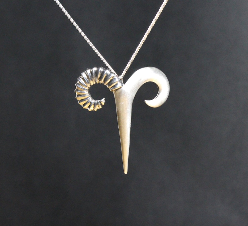 May include: A silver pendant necklace featuring a stylized ram's head design. The pendant has a detailed, curled horn and a smooth, pointed horn.