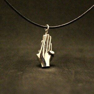 May include: A silver pendant necklace featuring a pair of hands clasped in prayer. The necklace is on a black leather cord.