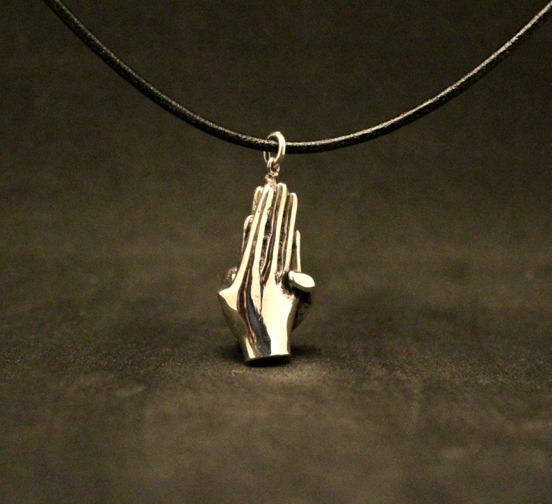 May include: A silver pendant necklace featuring a pair of hands clasped in prayer. The necklace is on a black leather cord.