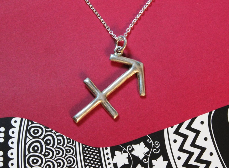 May include: A silver Sagittarius zodiac sign pendant on a silver chain. The pendant is hanging against a red background.