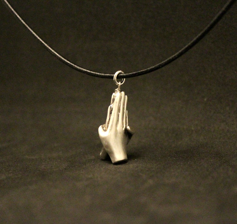 May include: A silver necklace with a pendant shaped like two hands clasped in prayer. The necklace is on a black leather cord.