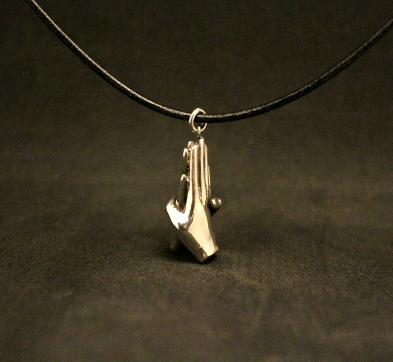 May include: A silver necklace with a pendant of two hands clasped together. The necklace is on a black cord.