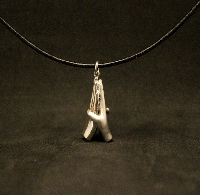May include: A silver necklace with a pendant of two hands clasped together in prayer. The necklace is on a black leather cord.
