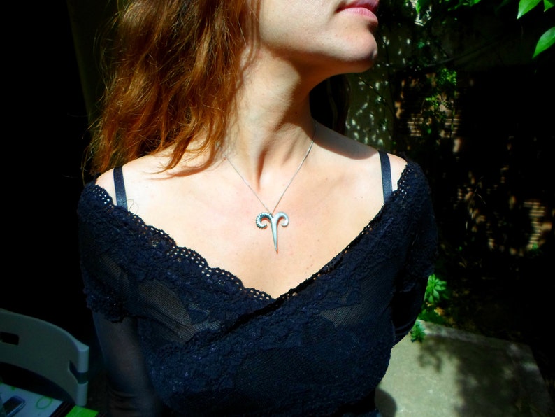May include: A woman wearing a black lace top with a silver ram pendant necklace.