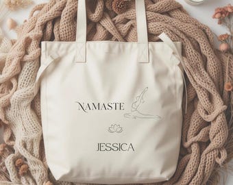 May include: Cream-colored tote bag with the word "NAMASTE" and the name "JESSICA" printed in black. A lotus flower and a yoga pose are also printed on the bag. The bag has two cream-colored handles.