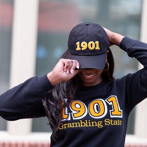 May include: Black baseball cap and sweatshirt with the number "1901" in gold lettering. The sweatshirt also has the words "Grambling State" below the number. The person is wearing gold hoop earrings.
