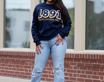 May include: A navy blue sweatshirt with the text "1891 North Carolina A&T" in gold and white. The sweatshirt is paired with light blue ripped jeans and white trainers. The person is standing in front of a brick wall.