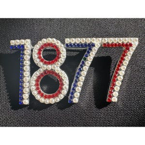 May include: A decorative pin featuring the number "1877". The numbers are outlined with small, round, white faux pearls. The "1" and "7" are accented with blue and red rhinestones, while the "8" is accented with red rhinestones.
